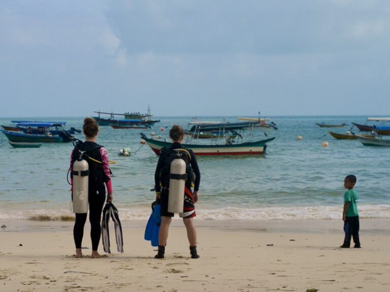Two scuba divers with Perhentian Marine Research Station wearing cylinders and holding their fins standing on a beach looking out towards the boats on the water. A small child looks at them from the right.