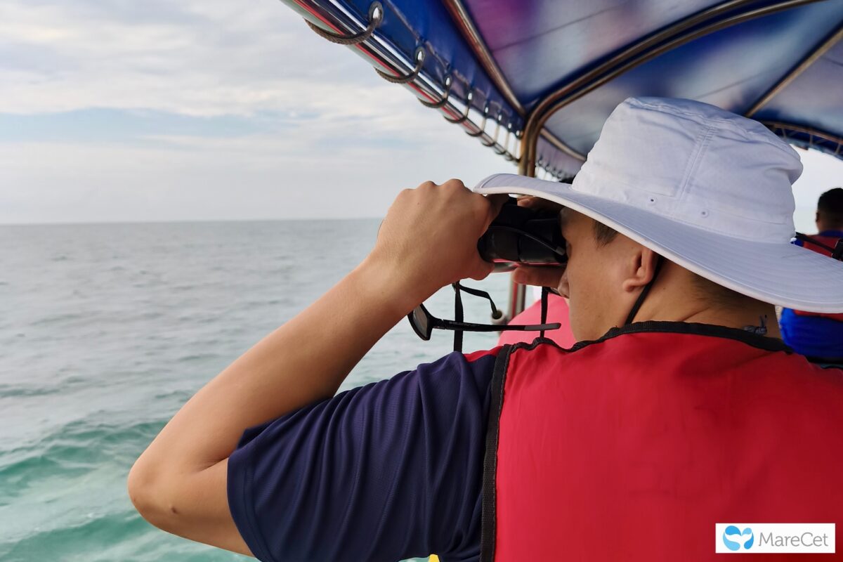 Back view of a man in a covered boat wearing a red life preserver and a white wide-brimmed hat. He has a pair of binoculars held up to his eyes looking out at sea for dugong.